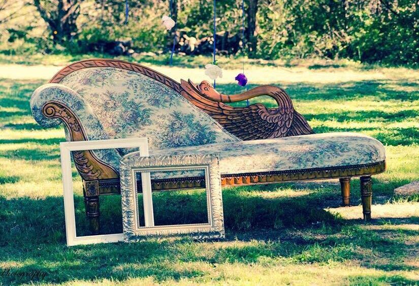 A blue and white chaise longue with a wood carved into the shape of a swan. It is sitting in a grassy field. Two white frames lean against it.